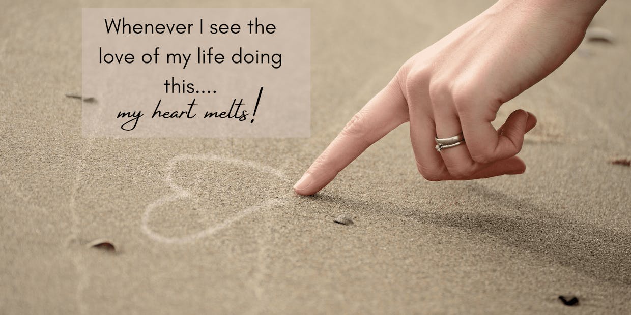 A lady draws a heart in the sand while wearing a white gold solitaire diamond ring and a plain white gold wedding band.